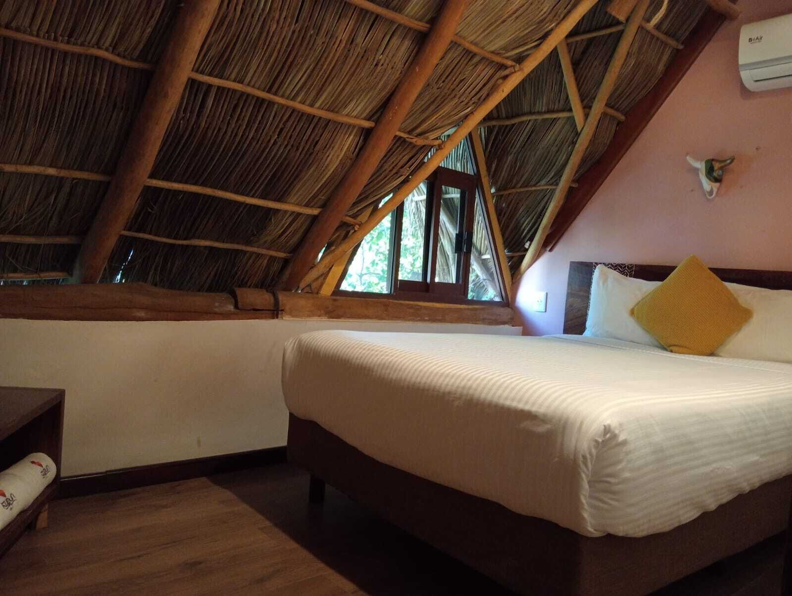 Un dormitorio con techo inclinado, cama con ropa de cama blanca, cabecera de madera y ventana con vista a la vegetación.