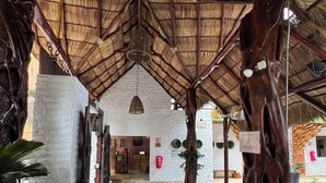 A covered walkway with a tiled floor, a reception area with a white wall and a wooden reception desk, and a pool area visible in the background. - Hotel Kupuri Sayulita (Sayulita)