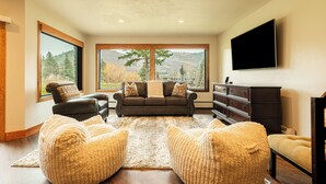 Ein Wohnzimmer mit einer Couch, Poufs, einem Flachbildfernseher und großen Fenstern mit Blick auf die Berge.