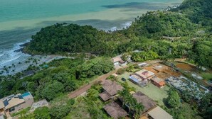 An aerial view of a coastal area with dense greenery, a beach, and several buildings with thatched roofs.