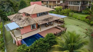 A two-story house with a swimming pool, surrounded by lush greenery and palm trees.