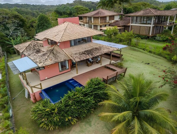 Une maison à deux étages avec une piscine, entourée d’une végétation luxuriante et de palmiers.