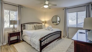 A bedroom with a bed, bedside tables, a ceiling fan, a mirror, and windows with blinds. - Sunset Rentals | Plantation Club 437 | Dog Friendly (Hilton Head Island)
