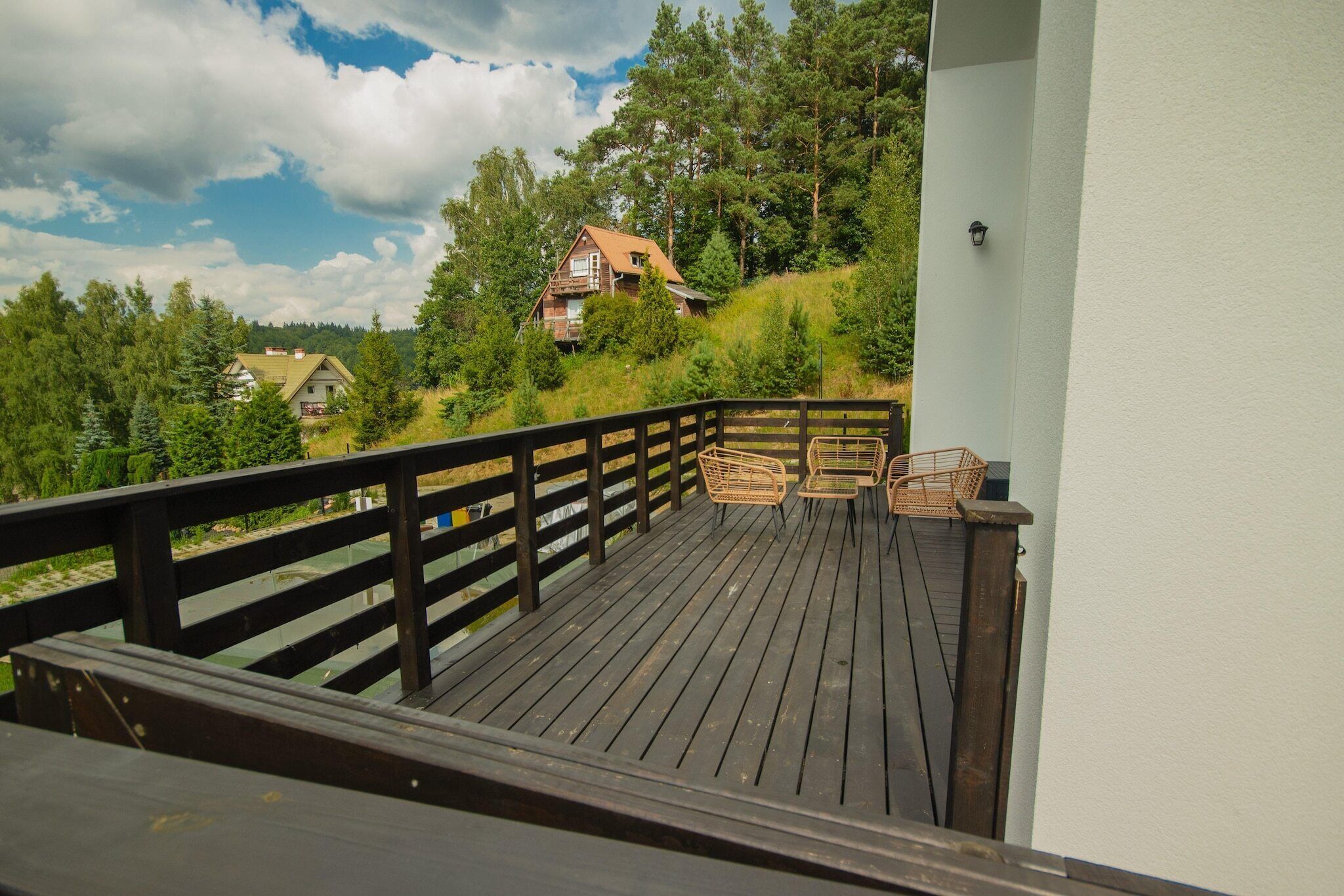 Eine Holzterrasse mit zwei Korbstühlen und einem kleinen Tisch mit Blick auf einen bewaldeten Hang und ein Haus.