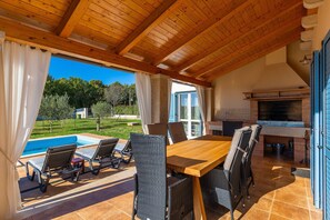 Un espace extérieur abrité, aménagé pour les repas, avec une piscine, un plafond en bois et une vue sur le jardin.