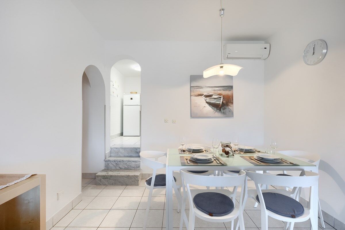 Un coin repas avec une table dressée pour quatre personnes, des chaises blanches avec des coussins, un tableau représentant un bateau accroché au mur et une horloge.