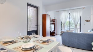 Un coin repas avec une table dressée pour deux personnes, un canapé et une bibliothèque, dans une pièce lumineuse dotée d’une grande fenêtre.