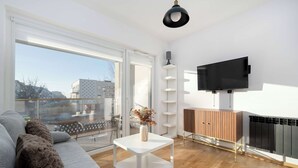 Un salon moderne avec un canapé, une table basse blanche, un téléviseur fixé au mur et une vue dégagée grâce à de grandes fenêtres.