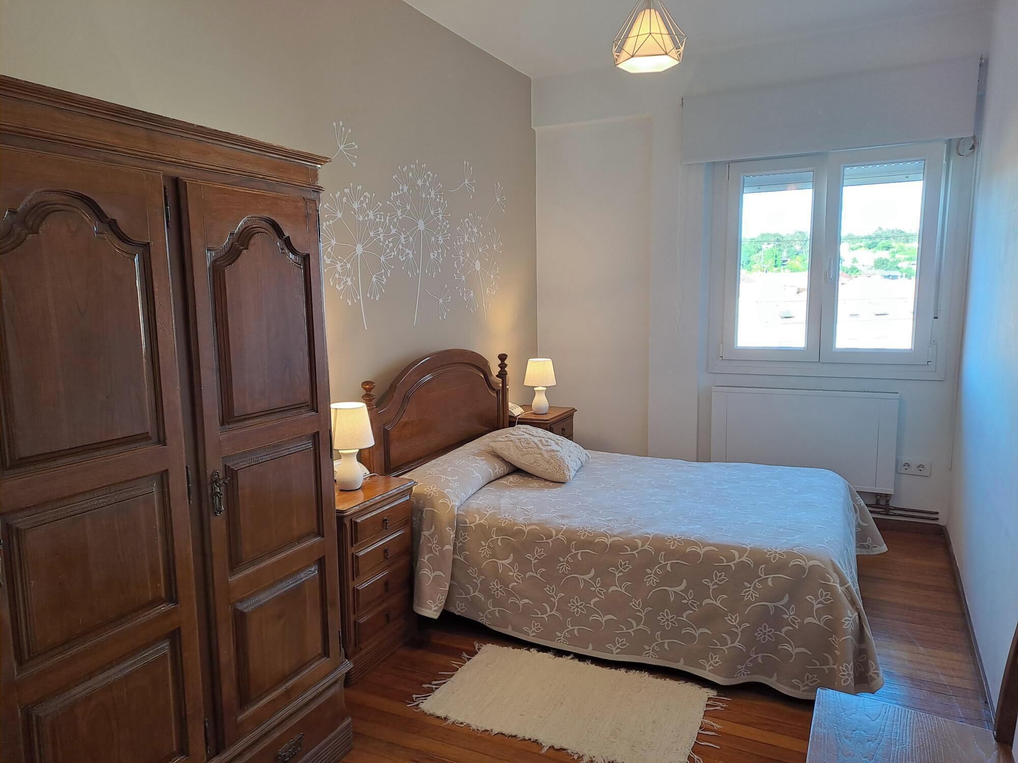 Ein Schlafzimmer mit Holzschrank, einem Bett mit Blumenmuster, Nachttischlampen, einem Fenster mit Aussicht und einer Deckenleuchte.