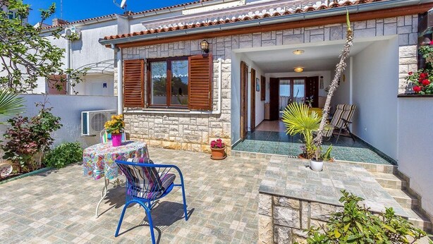 Ein Haus mit Natursteinfassade, Holzhörnern und einem Ziegeldach. Vor der Hauseingang befindet sich eine überdachte Terrasse mit Tisch und Stühlen sowie Blumentöpfen.