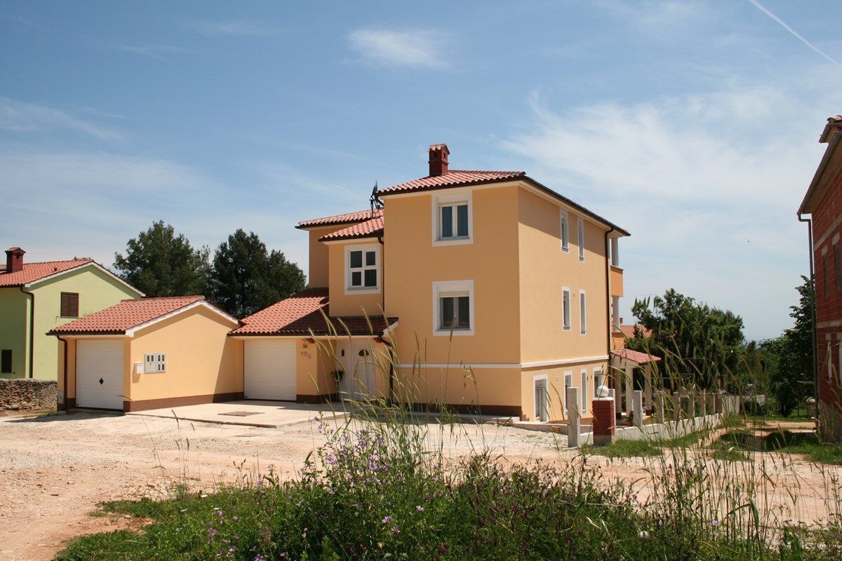 A two-story residential building with a red-tiled roof, a garage, and a small garden area with flowering plants.