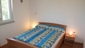 Una cama bien tendida con una colcha estampada, una mesita de noche con una lámpara y una ventana que da a un paisaje de árboles.