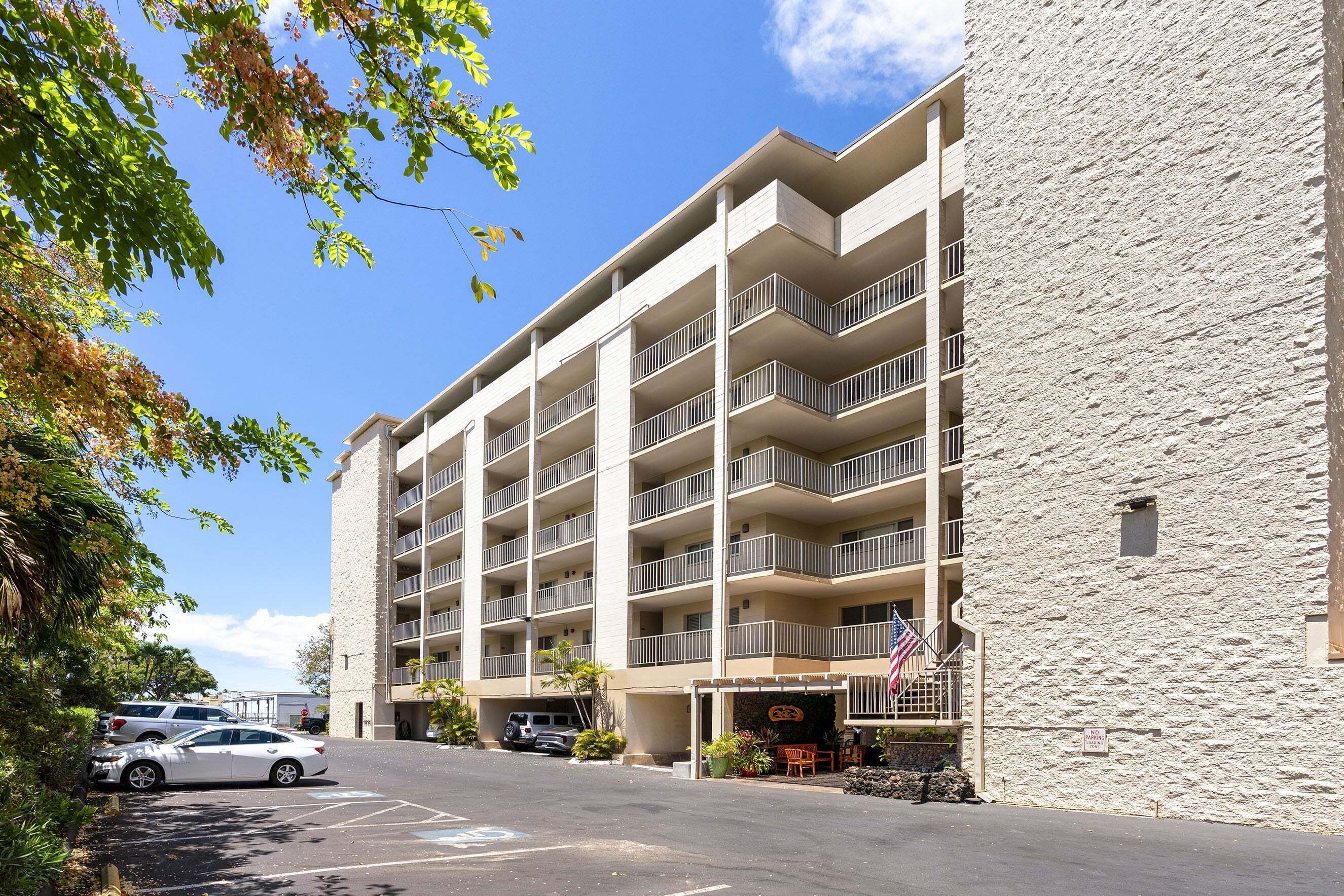Un edificio de varios pisos con balcones, estacionamiento y una pequeña zona exterior con plantas.