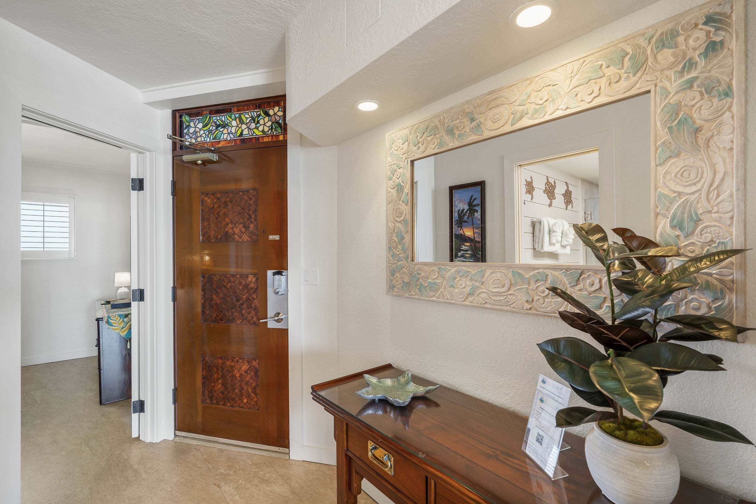A hotel reception area with a wooden door, a glass desk, a potted plant, and a mirror on the wall.