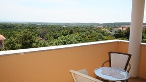 Uma varanda com vista para uma paisagem verde e edifícios. Há uma mesa e duas cadeiras.