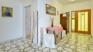 Un couloir avec un sol carrelé, une chaise blanche, une table recouverte d’une nappe rose et une porte avec un cadre en bois.