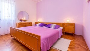Un dormitorio con cama de madera, ropa de cama morada, un espejo redondo, mesitas de noche con lámparas y suelo de madera.