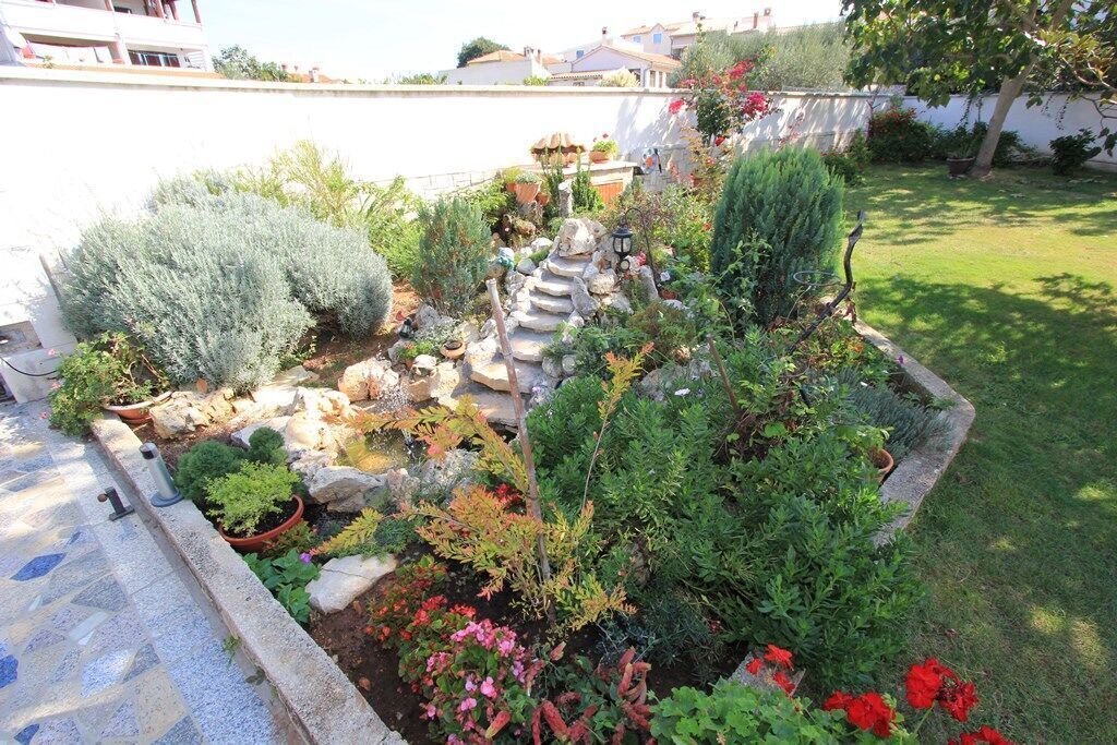Un giardino con diverse piante, una fontana in pietra e un vialetto lastricato.