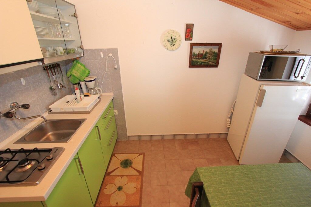 Cocina compacta con estufa a gas, lavamanos y armarios verdes. Hay un microondas y un refrigerador a la derecha, y un reloj y un cuadro en la pared.