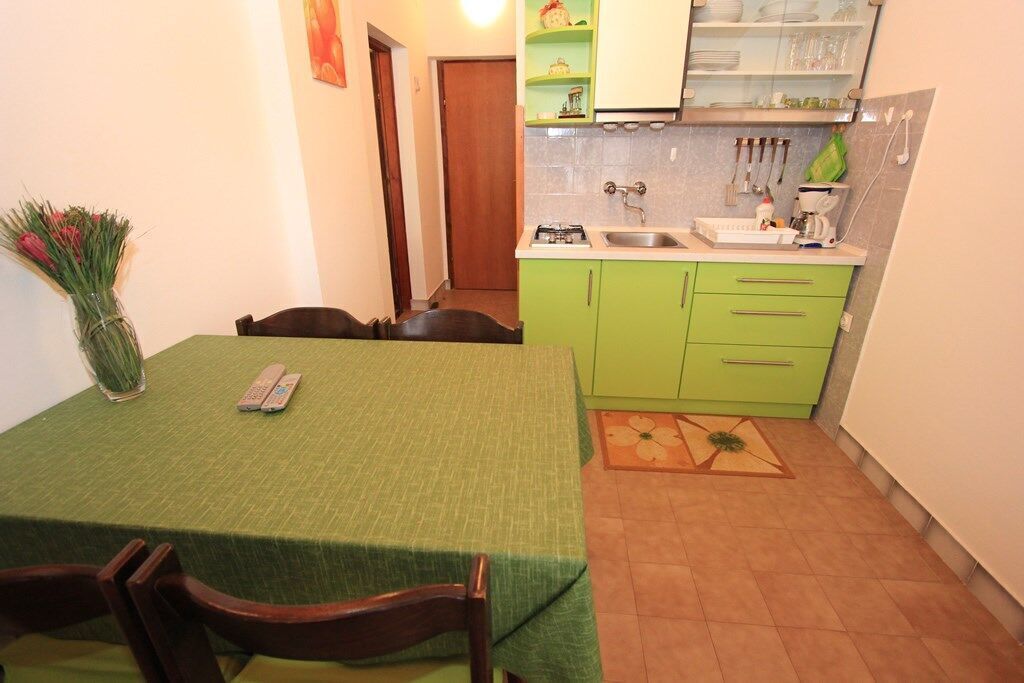 Uma cozinha compacta com armários verdes, uma mesa de jantar com cadeiras e um vaso com flores.