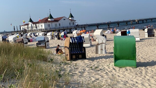 A beach with beach chairs and umbrellas, a white building with green roofs, and a pier extending into the water. - Apartment with Private Terrace, Shared Garden, and Wi-Fi in Heringsdorf (Heringsdorf)