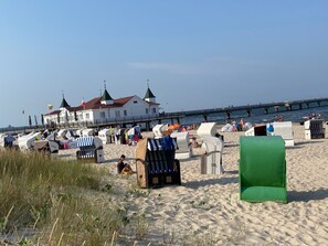 Een strand met ligbedden en parasols, een wit gebouw met groene daken, en een pier die het water in reikt.