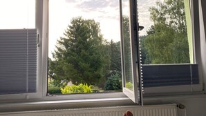 A room with a bed, a window with blinds, and a view of trees and a building outside. - Apartment with Private Terrace, Shared Garden, and Wi-Fi in Heringsdorf (Heringsdorf)