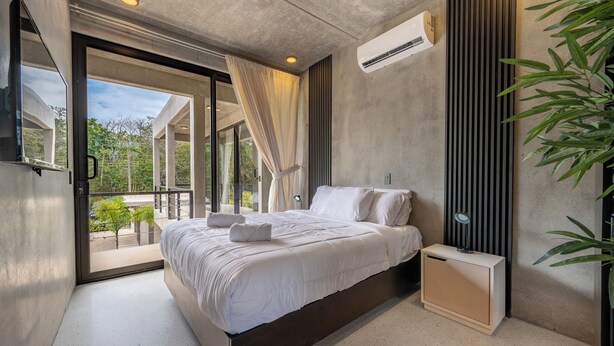 A modern bedroom with a large bed, a bedside table, a built-in bench, and a view of greenery outside. - NELBLU FLAMINGO SUITES (Playa Flamingo)