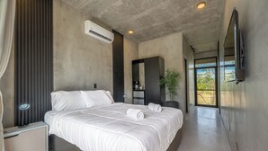 A modern bedroom with a large bed, a bedside table, an air conditioning unit, a mirror, and a view of the outdoors. - NELBLU FLAMINGO SUITES (Playa Flamingo)