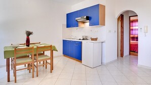 A compact kitchen with blue cabinets, a white countertop, and a dining area with wooden chairs and a green tablecloth. - Apartments Ulika (16031-A4) - Medulin (Medulin)
