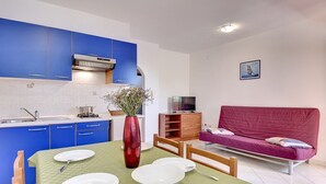 A compact living space with a kitchenette, a dining table set with plates and cutlery, a red vase with flowers, a sofa, and a television. - Apartments Ulika (16031-A4) - Medulin (Medulin)