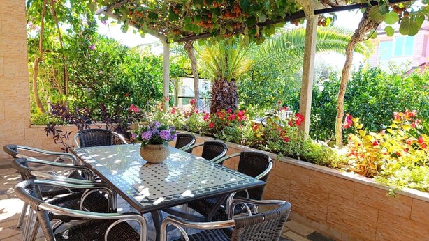 Un patio al aire libre con una mesa de vidrio y sillas de mimbre, rodeado de abundante vegetación y plantas en flor.