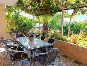 Um pátio externo com mesa de vidro e cadeiras de vime, cercado por vegetação exuberante e plantas floridas.