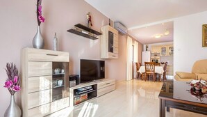 Una sala de estar moderna con un televisor de pantalla plana, armarios de madera, una zona de comedor con mesa y sillas, y una cocina al fondo.