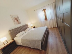 Quarto com cama, mesas de cabeceira, abajur, janela com cortinas e guarda-roupa.