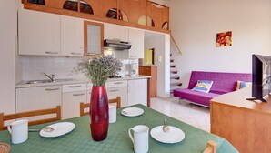 Eine kleine Küche mit weißen Schränken, ein Esstisch mit grüner Tischdecke und ein violettes Sofa im Wohnbereich.