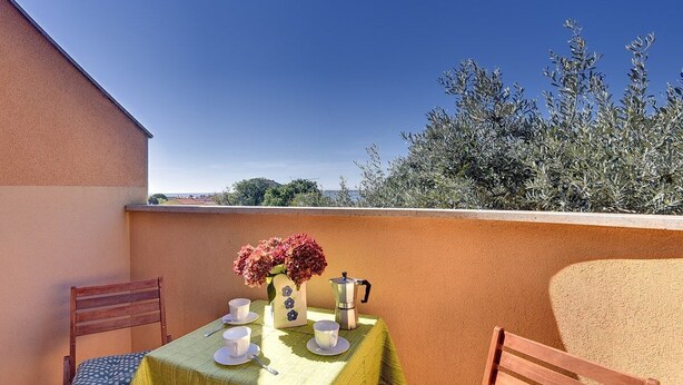 Ein Tisch mit Tassen, Untertassen, einer Blumenvase und einer Kaffeekanne auf einem Balkon mit Blick auf Bäume und ein Gebäude.