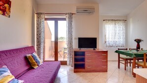 Ein Wohnzimmer mit einem violetten Sofa, einem Holz-TV-Schrank, einem Esstisch mit Stühlen und einer Glasschiebetür, die zu einem Außenbereich führt.