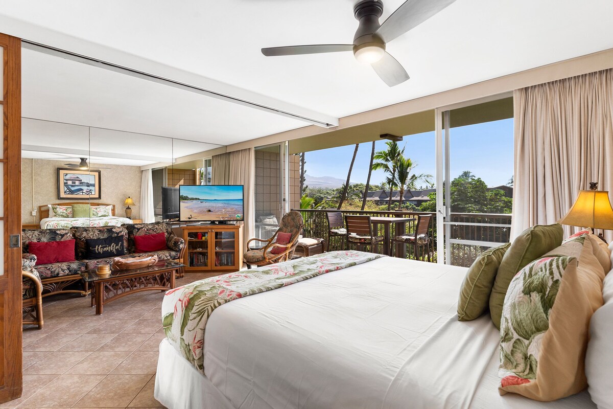 A hotel room with a large bed, a sofa, a dining table, and a balcony with a view of palm trees and mountains.