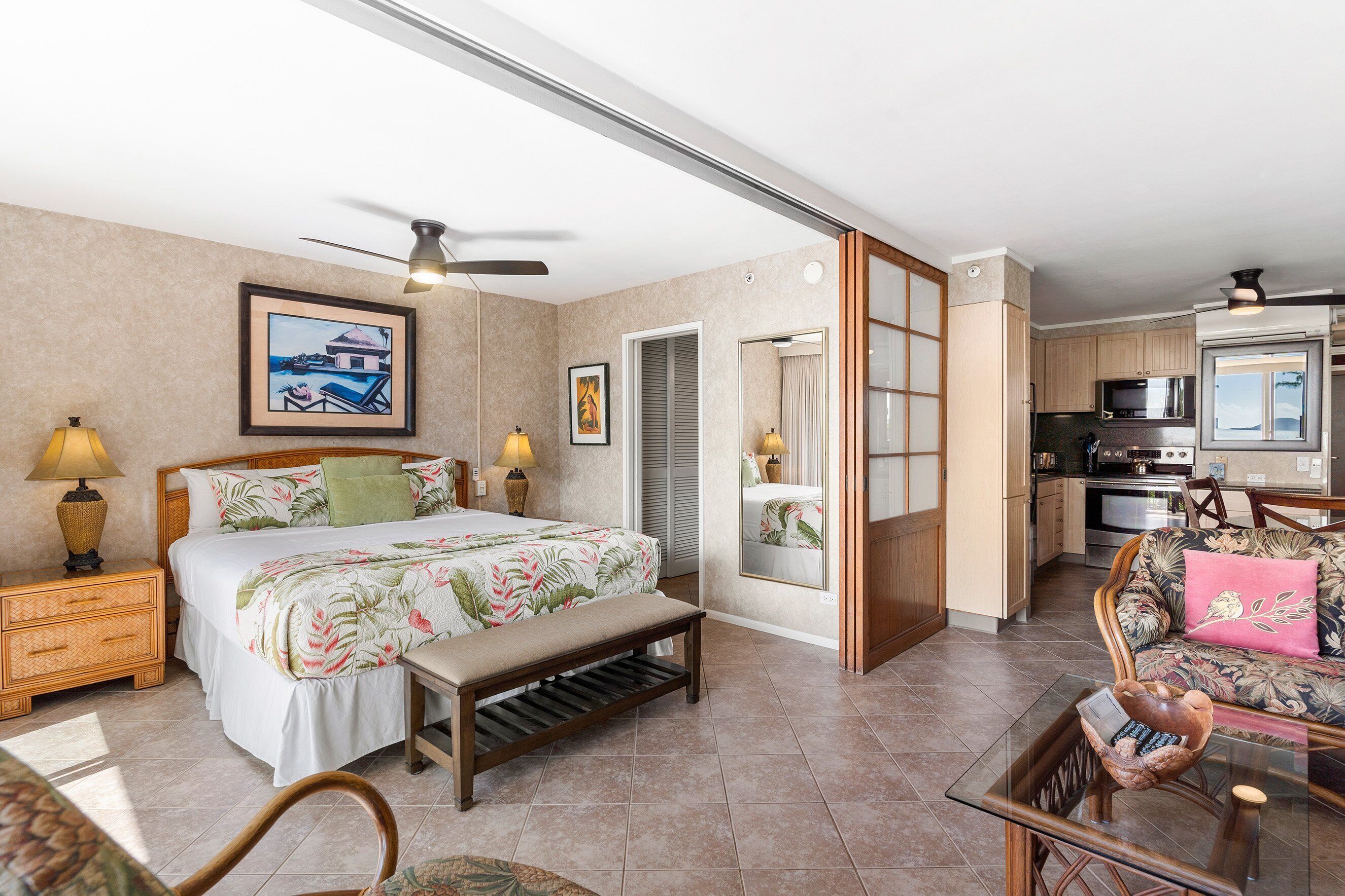 A bedroom with a bed, bedside tables, a bench, and a view of the kitchen.