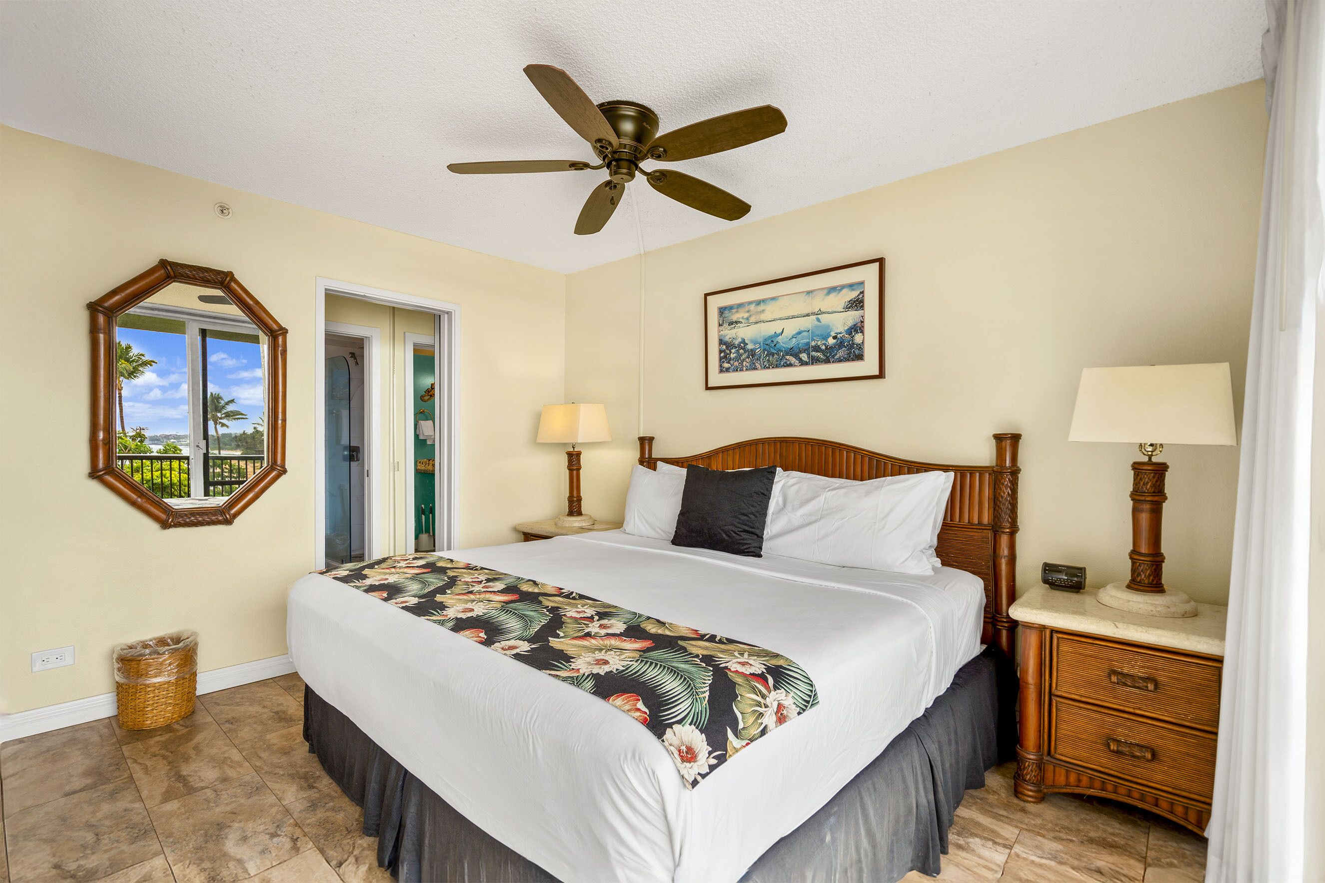 A hotel room with a bed, a ceiling fan, a nightstand with a lamp, a framed picture on the wall, and a door leading to a balcony.