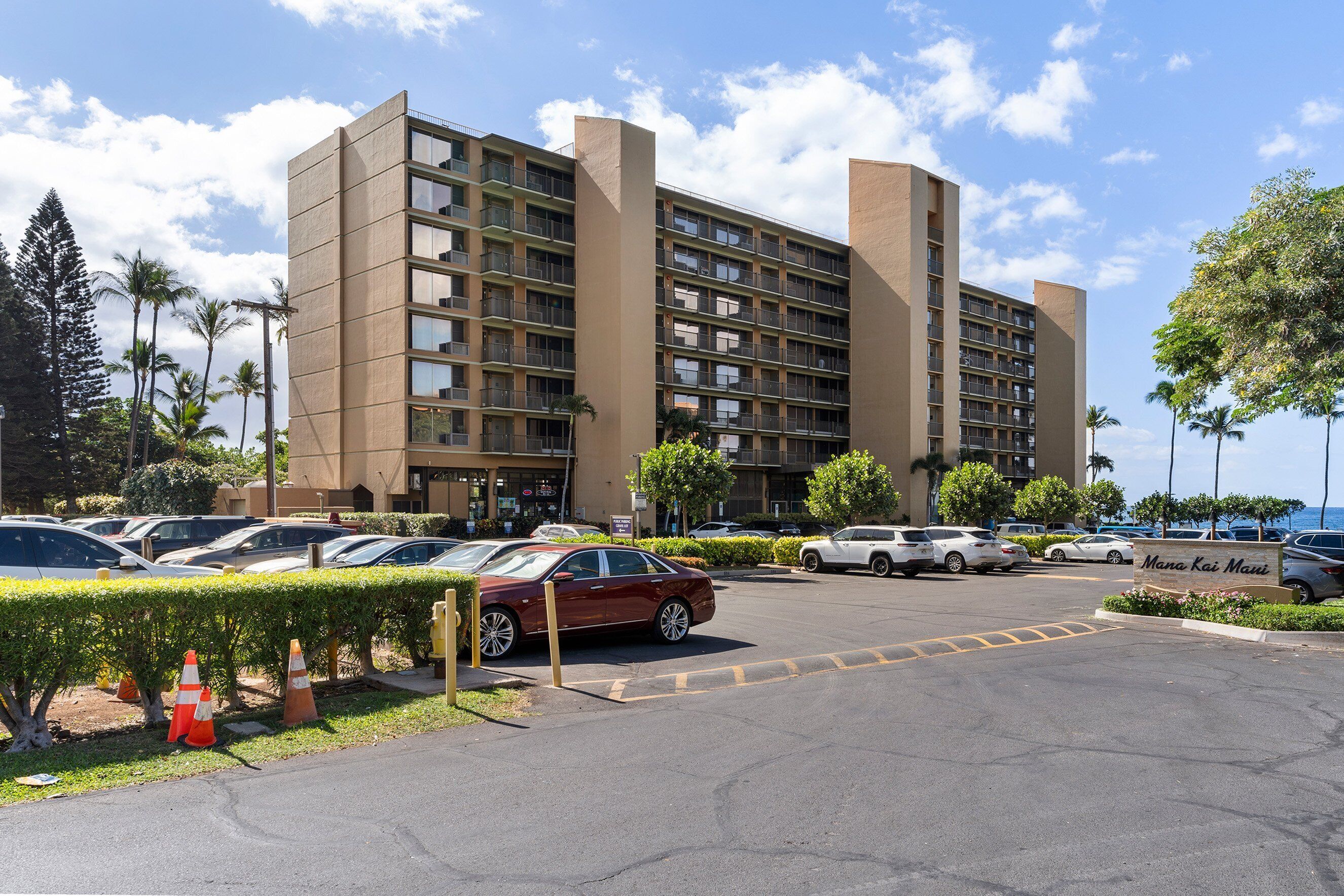 A multi-story hotel with a parking lot, several cars, and a sign that reads "Mana Kai Maui".