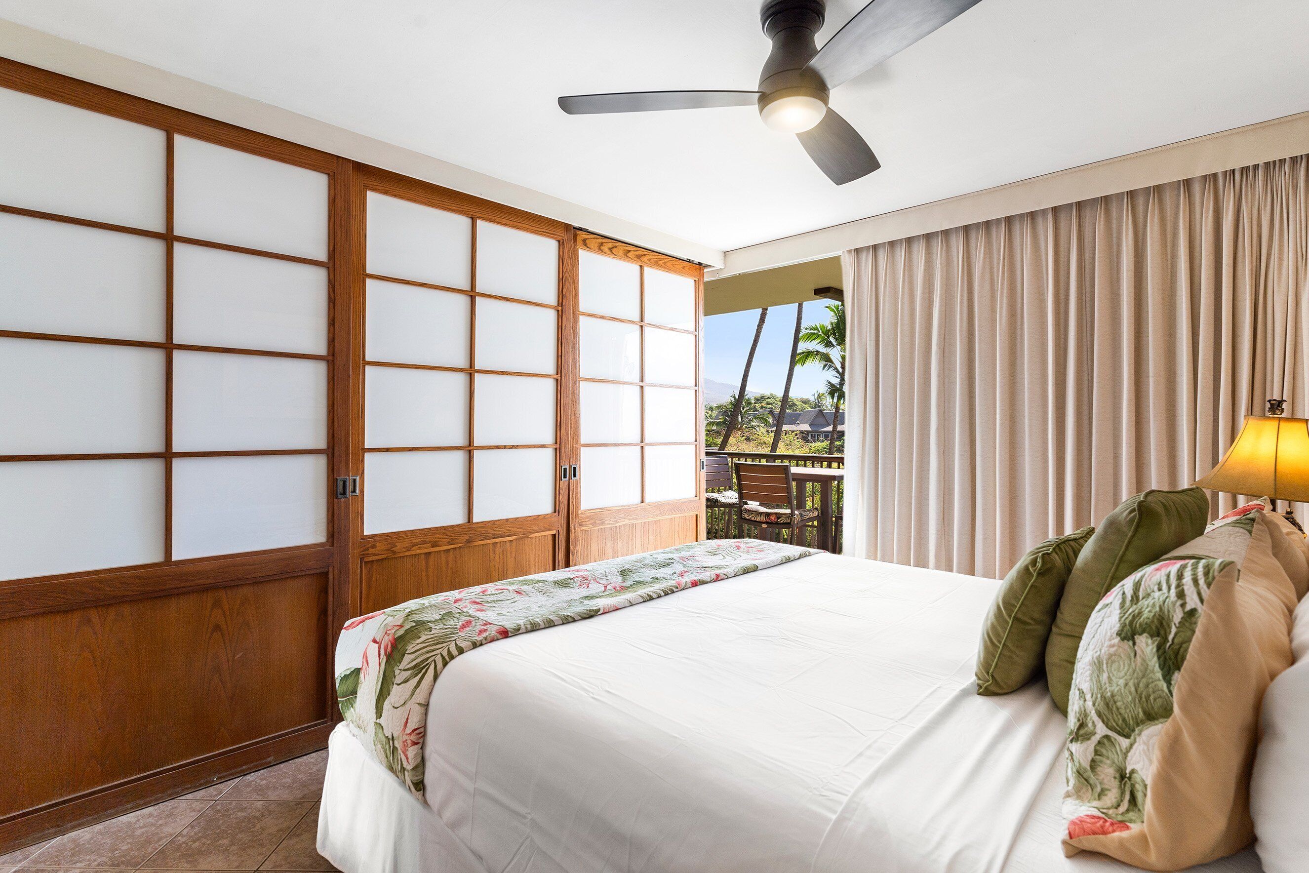 Habitación de hotel con una cama grande, ventilador de techo y vistas a palmeras, además de una puerta corredera.