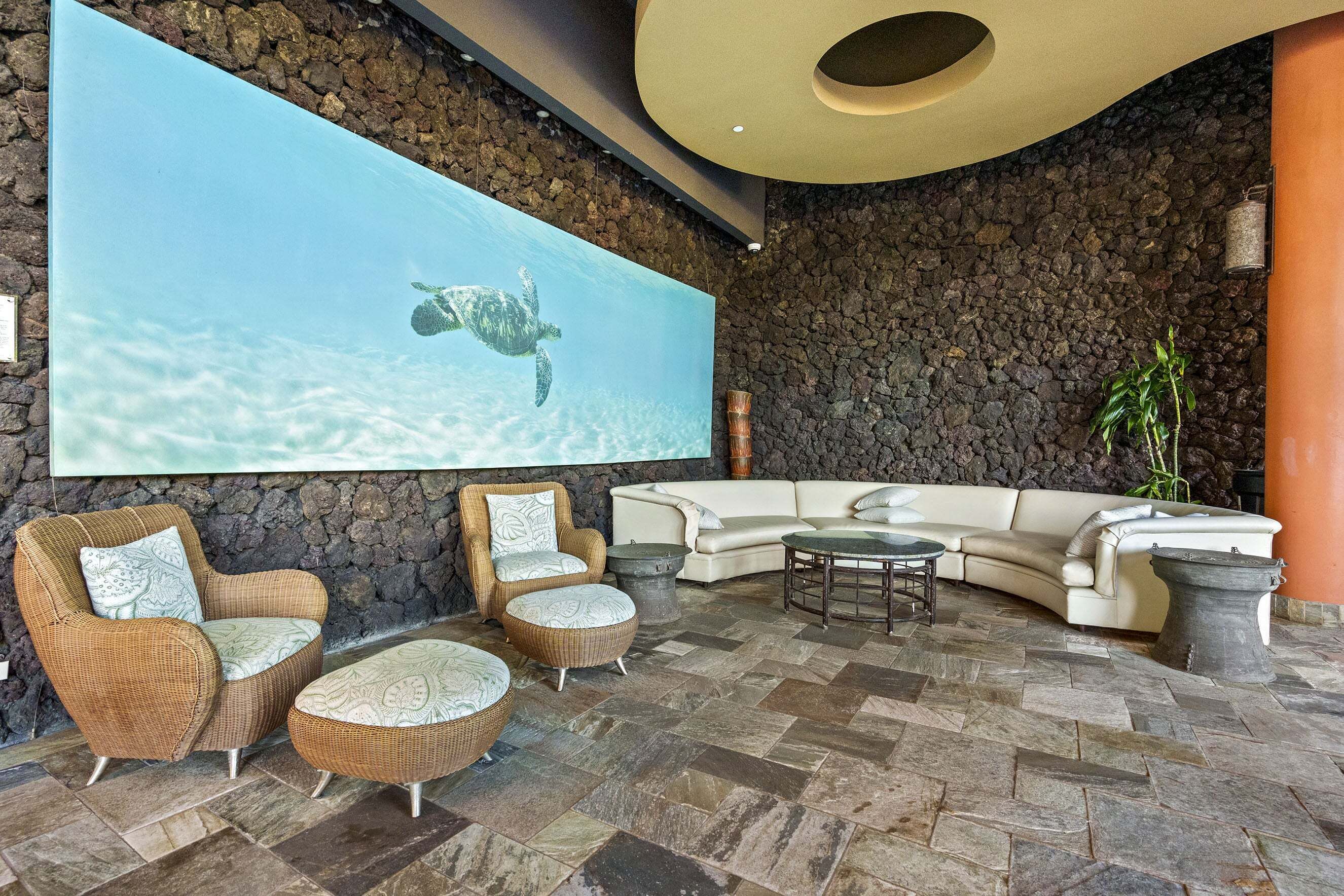 A lounge area with a stone wall, a large screen displaying a turtle, wicker furniture, and a stone floor.