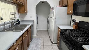 Una cocina con gabinetes de madera, un lavamanos de acero inoxidable, un refrigerador blanco, una estufa negra y piso de baldosas.