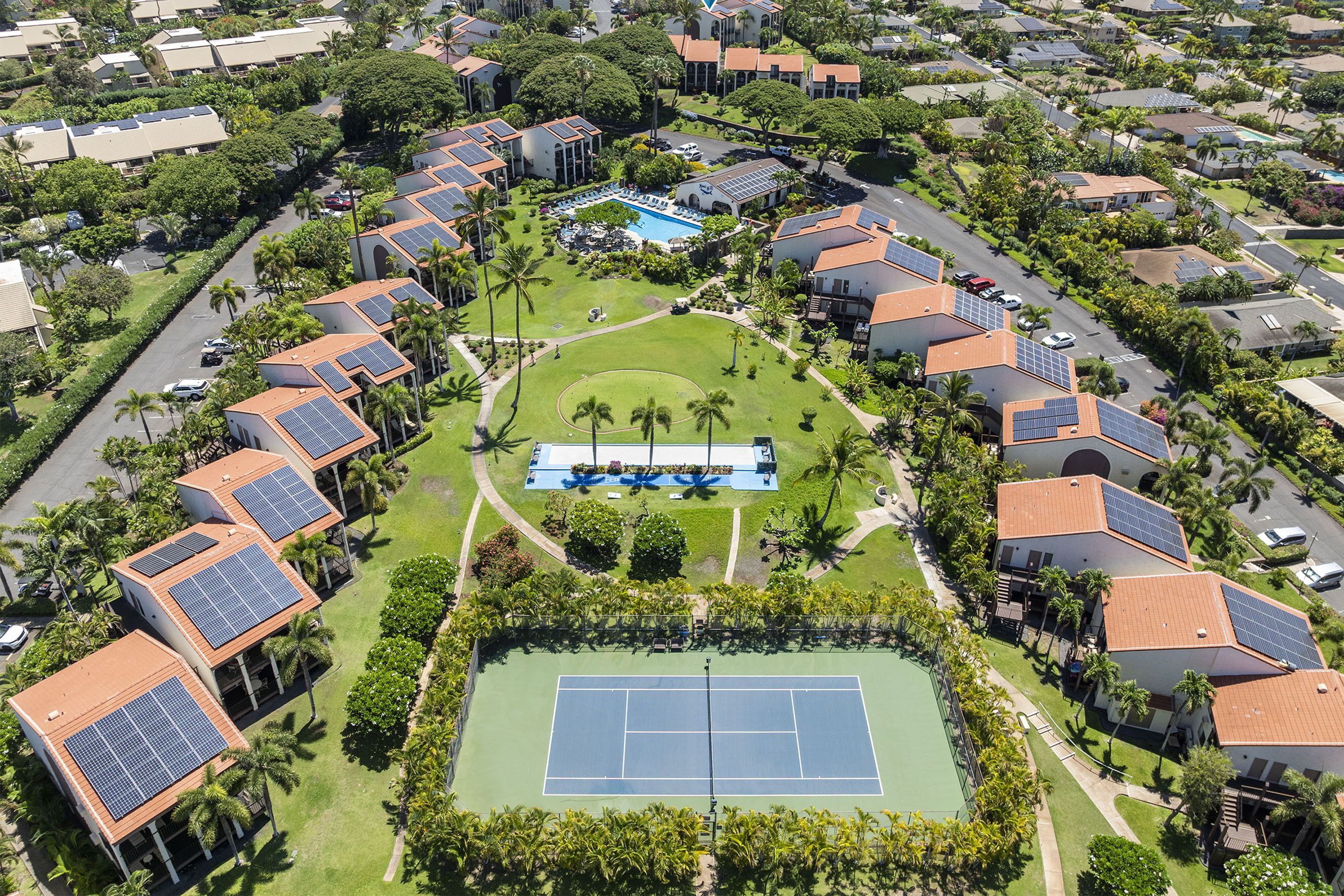 An aerial view of a residential area with solar panels on buildings, a tennis court, and a swimming pool.