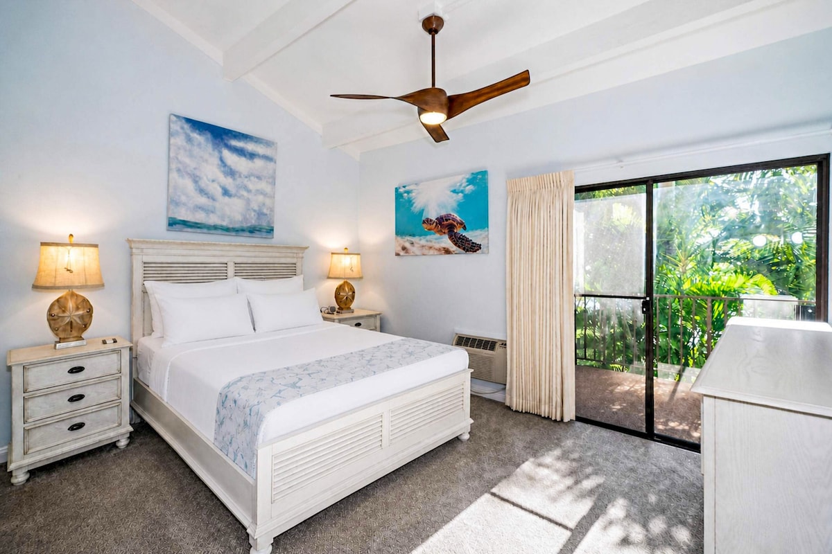 A bedroom with a bed, two bedside tables, a ceiling fan, and a view of greenery through a sliding glass door.