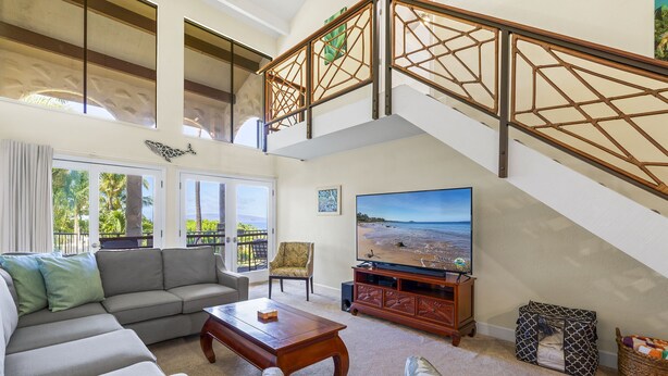 A spacious living room with a large flat-screen TV, a sofa, a coffee table, and a staircase with a decorative railing. - Maui Hill (Kihei)
