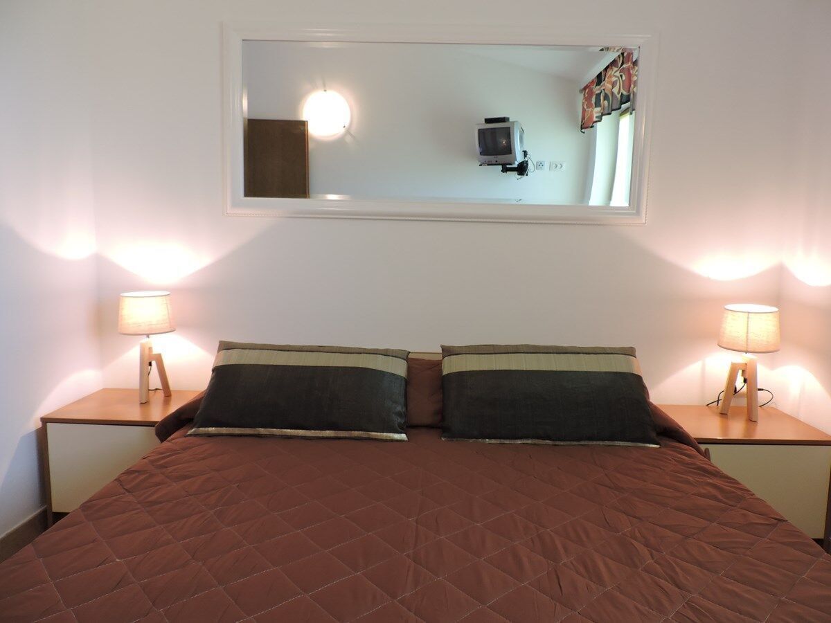 Ein Doppelbett mit einer braunen Tagesdecke, zwei Nachttische mit Lampen sowie ein Spiegel über dem Bett, der einen Fernseher und Vorhänge reflektiert.