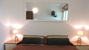 Cama doble con colcha marrón, dos mesitas de noche con lámparas y un espejo sobre la cama que refleja un televisor y cortinas.